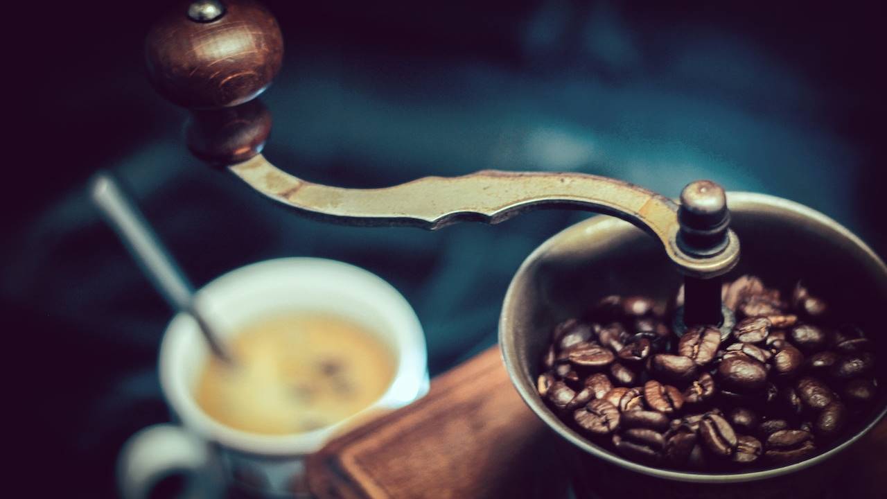Molinillo de café manual con grnos en su interior y taza de café en el fondo